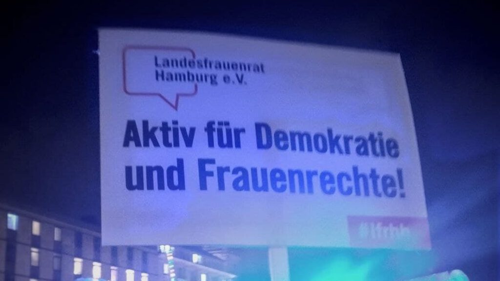 Foto eines Demoschildes in bläulichem Licht in städtischer Umgebung mit der Aufschrift: "Aktiv für Demokratie und Frauenrechte!"