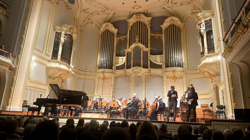 Aufnahme der Bühne des Großen Saals in der Laeiszhalle, zwei Frauen halten eine Rede, das Orchester wartet