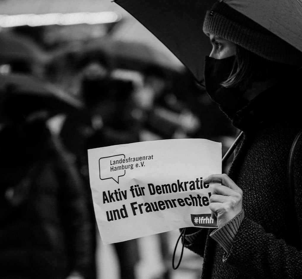 Schwarz-weiß Foto einer Person, die ein Schild in der Hand hält mit der Aufschrift: „Aktiv für Demokratie und Frauenrechte!“