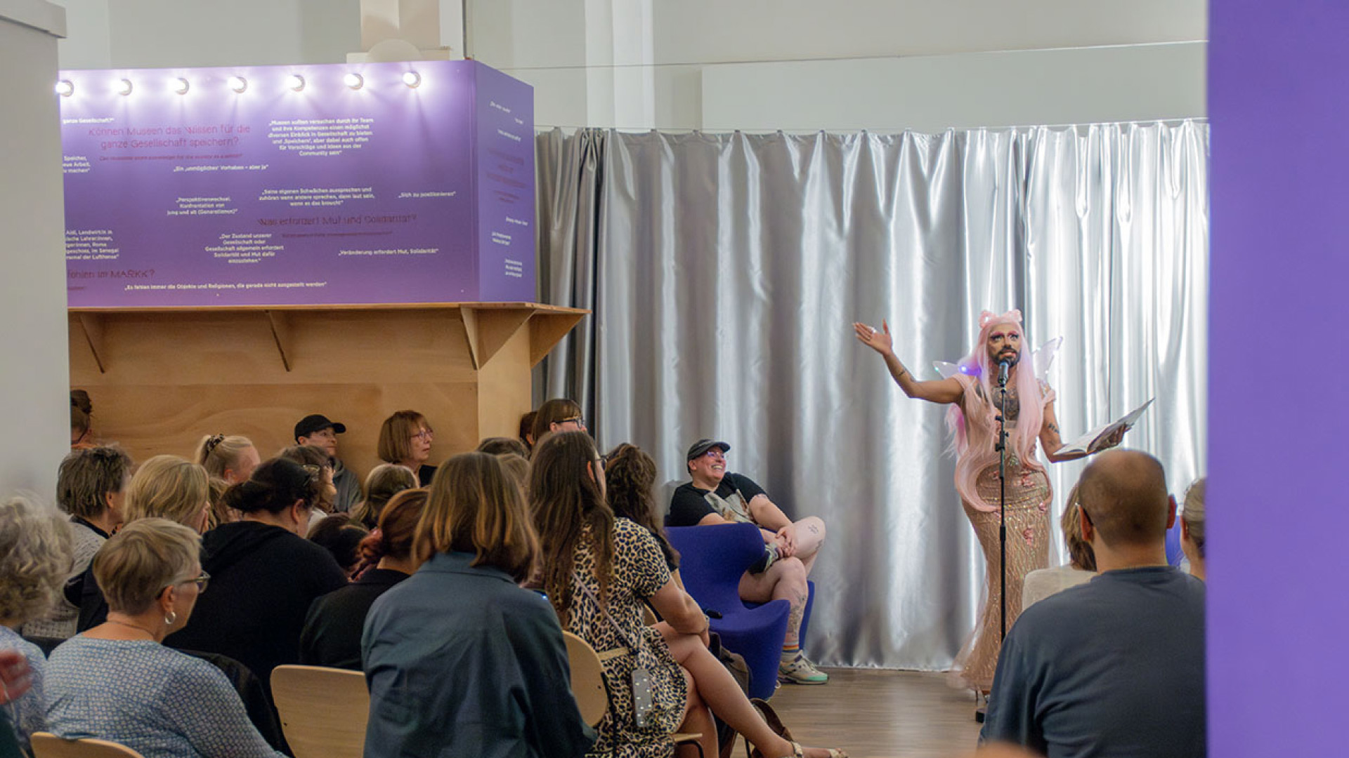 Drag Queen Lady Sasha steht vor einem Publikum am Mikrofon und macht eine theatralische Geste in die Luft, während sie aus einem Kinderbuch vorliest.